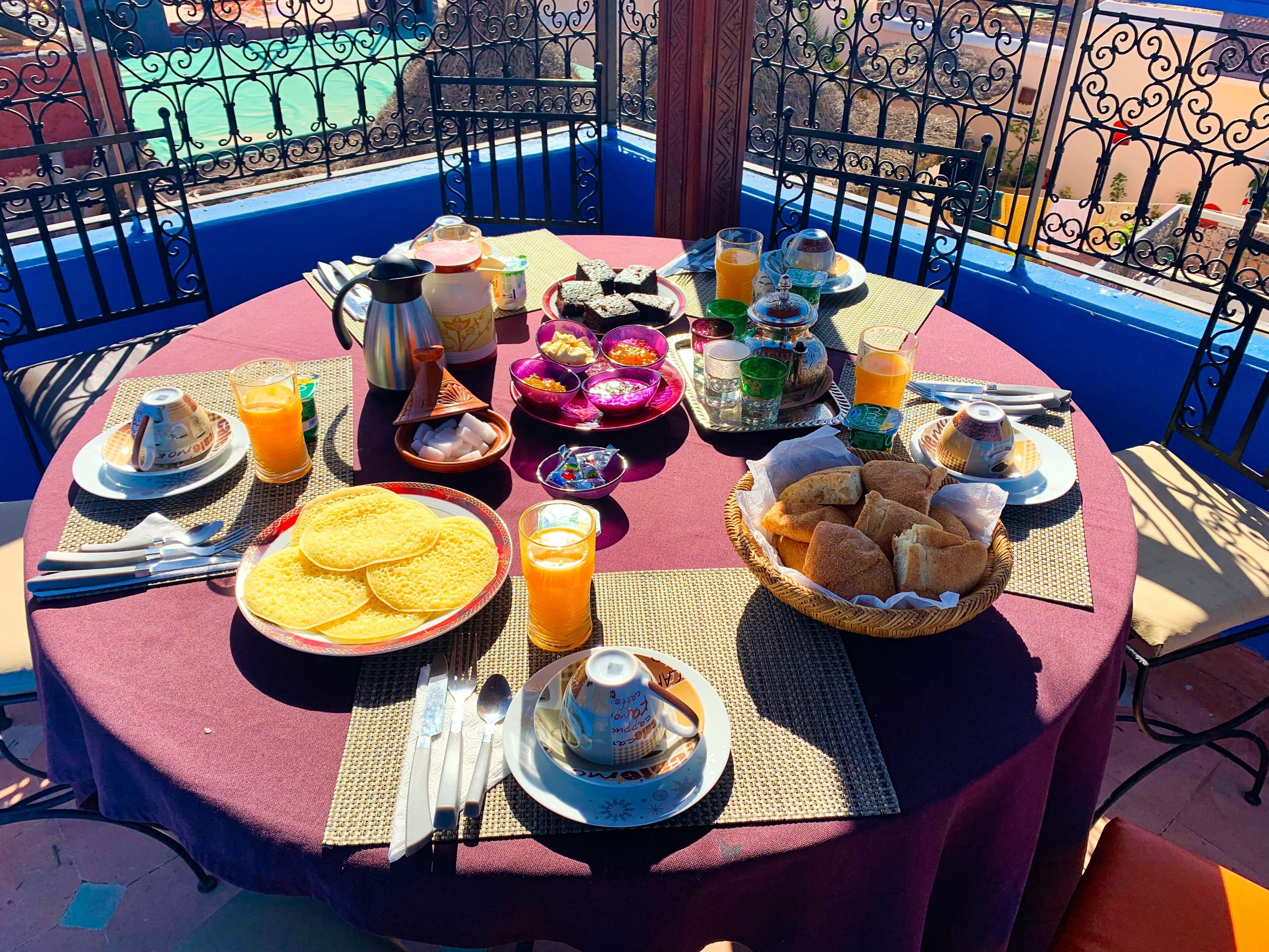A round table set with breakfast items, including bread, eggs, and beverages, on a balcony with a blue railing.