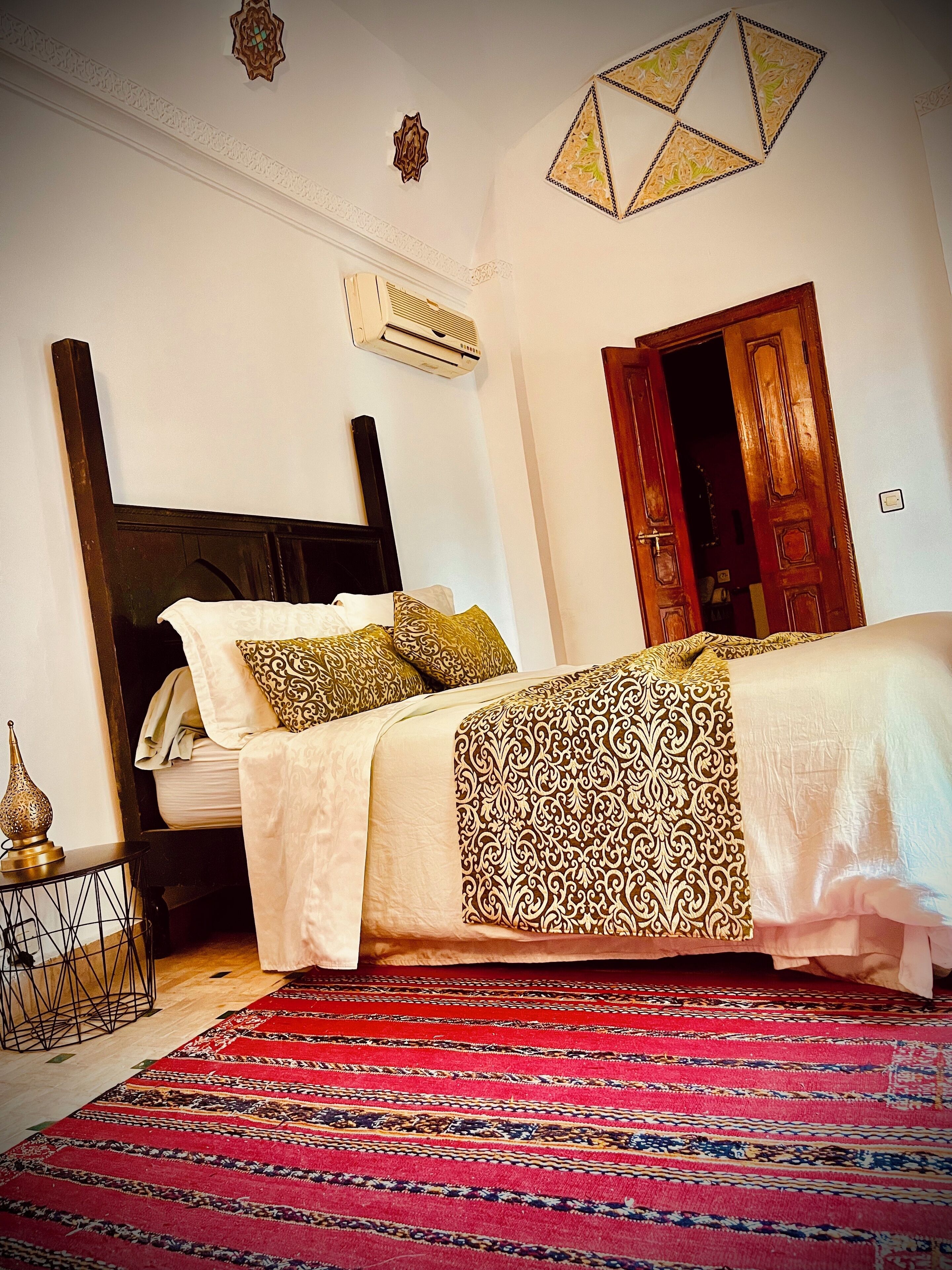 Quarto com uma cama, tapete vermelho estampado, almofadas decorativas e uma porta de madeira.