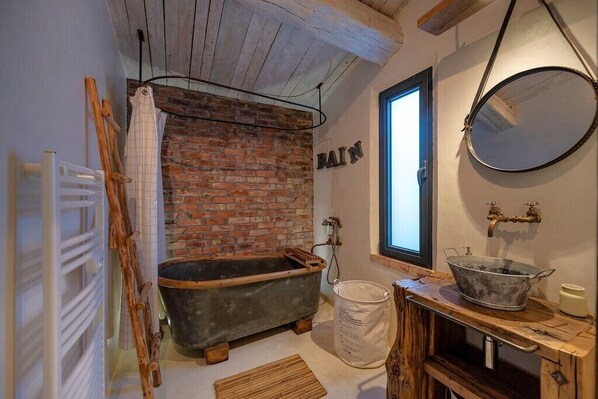A rustic bathroom with a brick wall, an antique bathtub, a wooden vanity, and a large mirror.