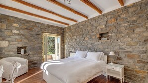 A bedroom with a stone wall, wooden beams, a bed, a nightstand, a chair, and a window. - Beachfront villa with stunning sea view . (Paros)