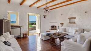 A spacious living room with white furniture, wooden beams, a fireplace, and a view of the sea through a window. - Beachfront villa with stunning sea view . (Paros)