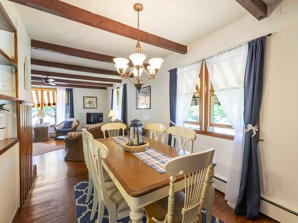 Un espace repas comprenant une table en bois, des chaises blanches, un lustre et une vue dégagée sur l’extérieur grâce à de grandes fenêtres.