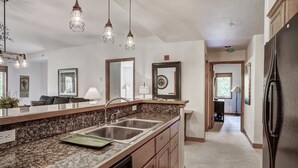 A kitchen with granite countertops, stainless steel sinks, and a dishwasher. There are pendant lights above the sink and a refrigerator on the right.