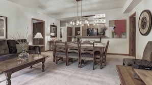 A spacious living room with a large wooden dining table surrounded by chairs, a kitchen area with wooden cabinets, and a dining area with a chandelier.