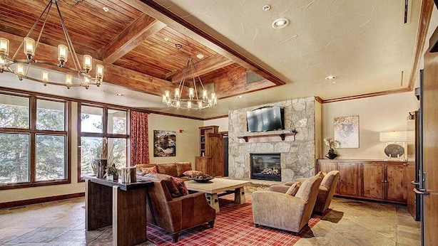 A spacious living room with a stone fireplace, wooden ceiling beams, and a large window overlooking a forest.