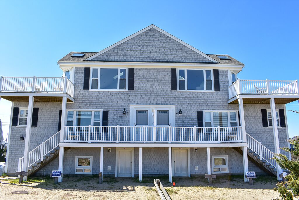 A two-story beachfront house with a balcony, a covered porch, and a sandy front yard.