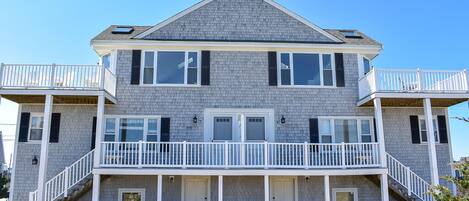 A two-story beachfront house with a balcony, a covered porch, and a sandy front yard.