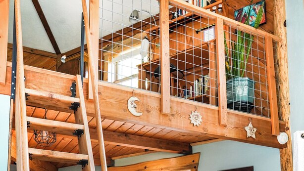 A wooden loft bedroom with a wooden ladder, a window with a view, and a kitchen area below.