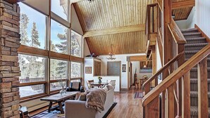 A spacious living area with a high ceiling, wooden staircase, and large windows offering a view of trees. - Breck Cabin, Ten Mile Range Views, Wrap Deck, Fireplaces, Sleeps 6 (Breckenridge)