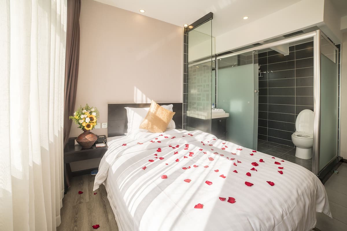A hotel room with a bed, a nightstand with a vase of flowers, a glass shower enclosure, and a toilet.