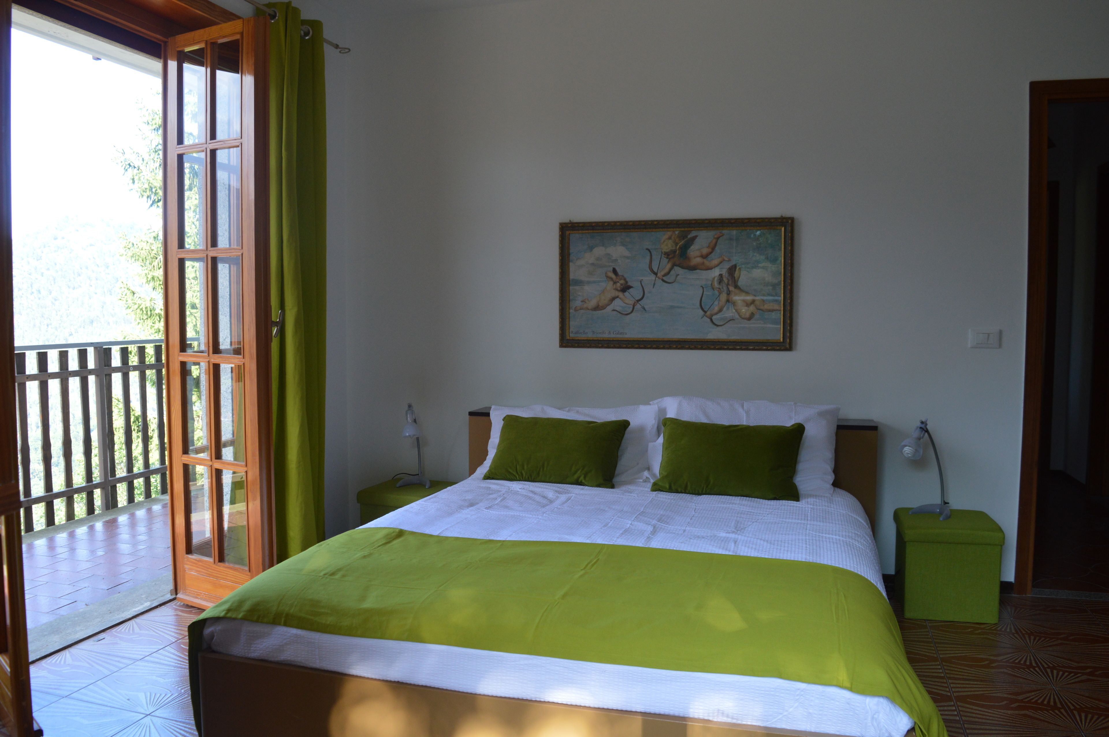 A bedroom with a bed, green pillows, a painting on the wall, and a view through a glass door.