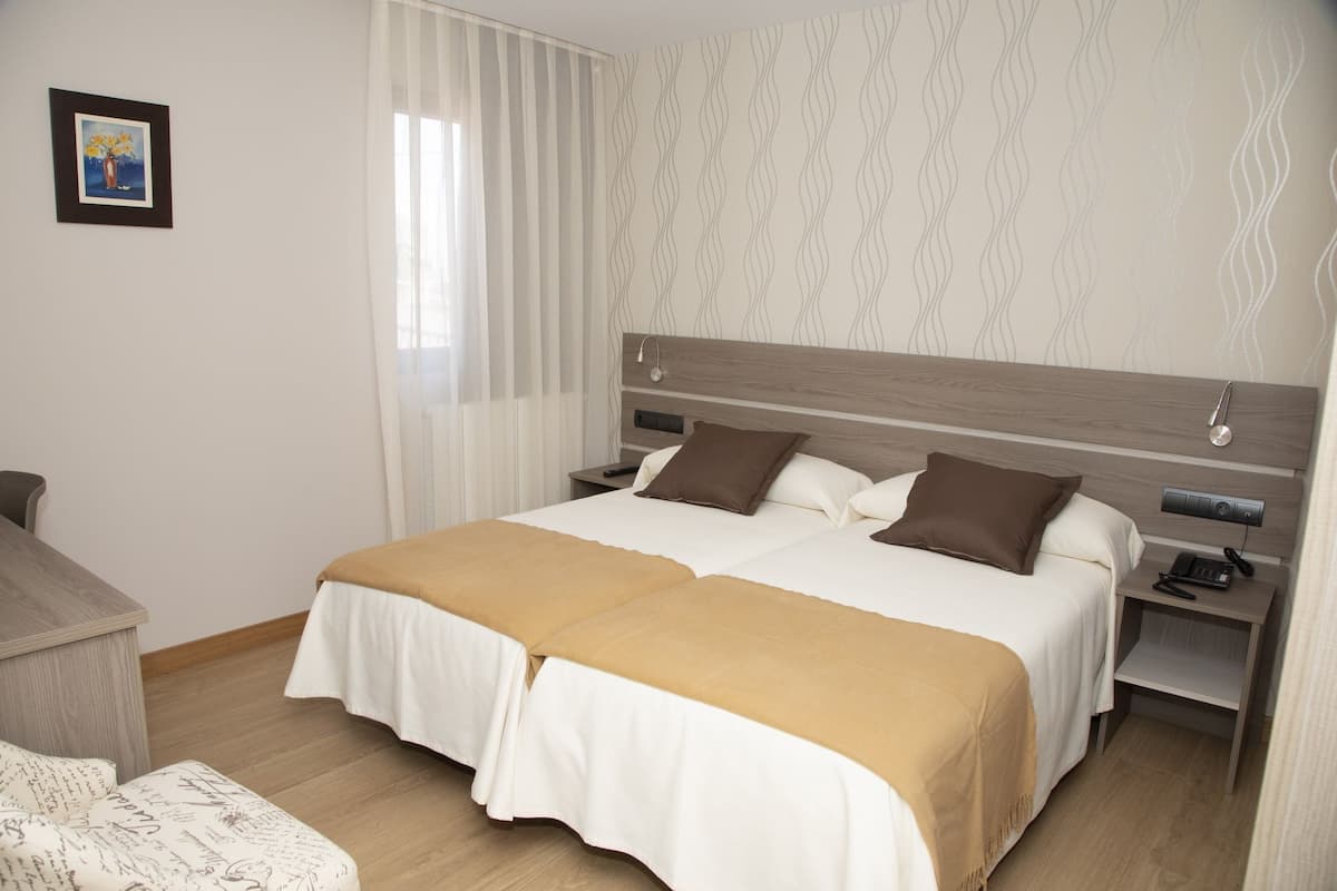 A double bed with two pillows, a bedside table with a phone, and a window with blinds.