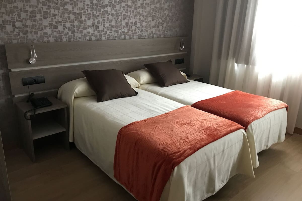 A double bed with a white and orange bedspread, a nightstand with a phone, and a headboard with a small shelf.