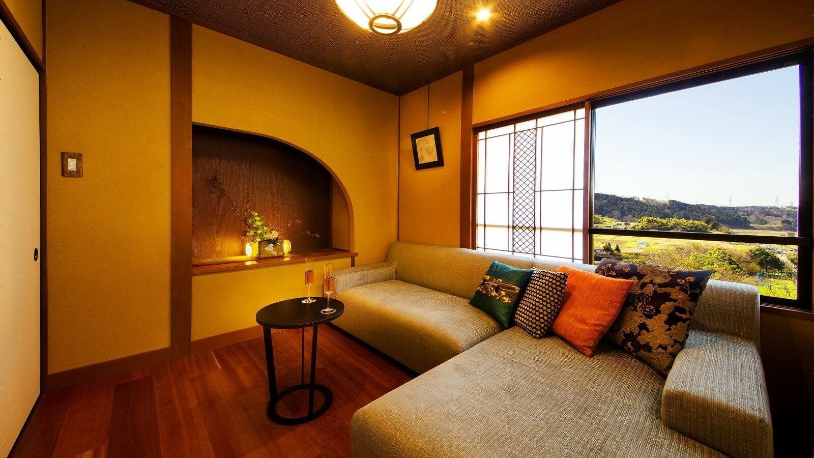 A room with a sofa, a small table with glasses, a window with a view, and a decorative shelf.