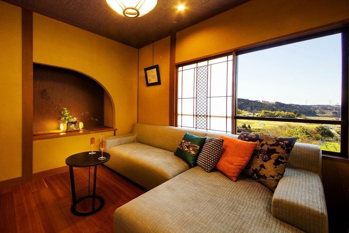 A room with a sofa, a small table with glasses, a window with a view, and a decorative shelf.