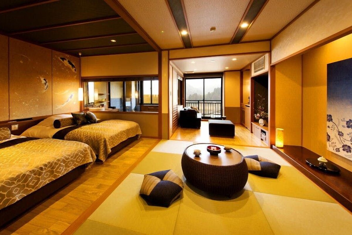 A traditional Japanese-style hotel room with tatami flooring, two beds, a tatami mat seating area, and a large window with a view.