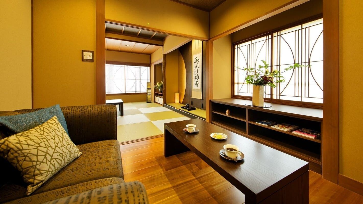 A traditional Japanese room with tatami flooring, a low wooden table with cups, a sofa with cushions, and a window with a geometric pattern.