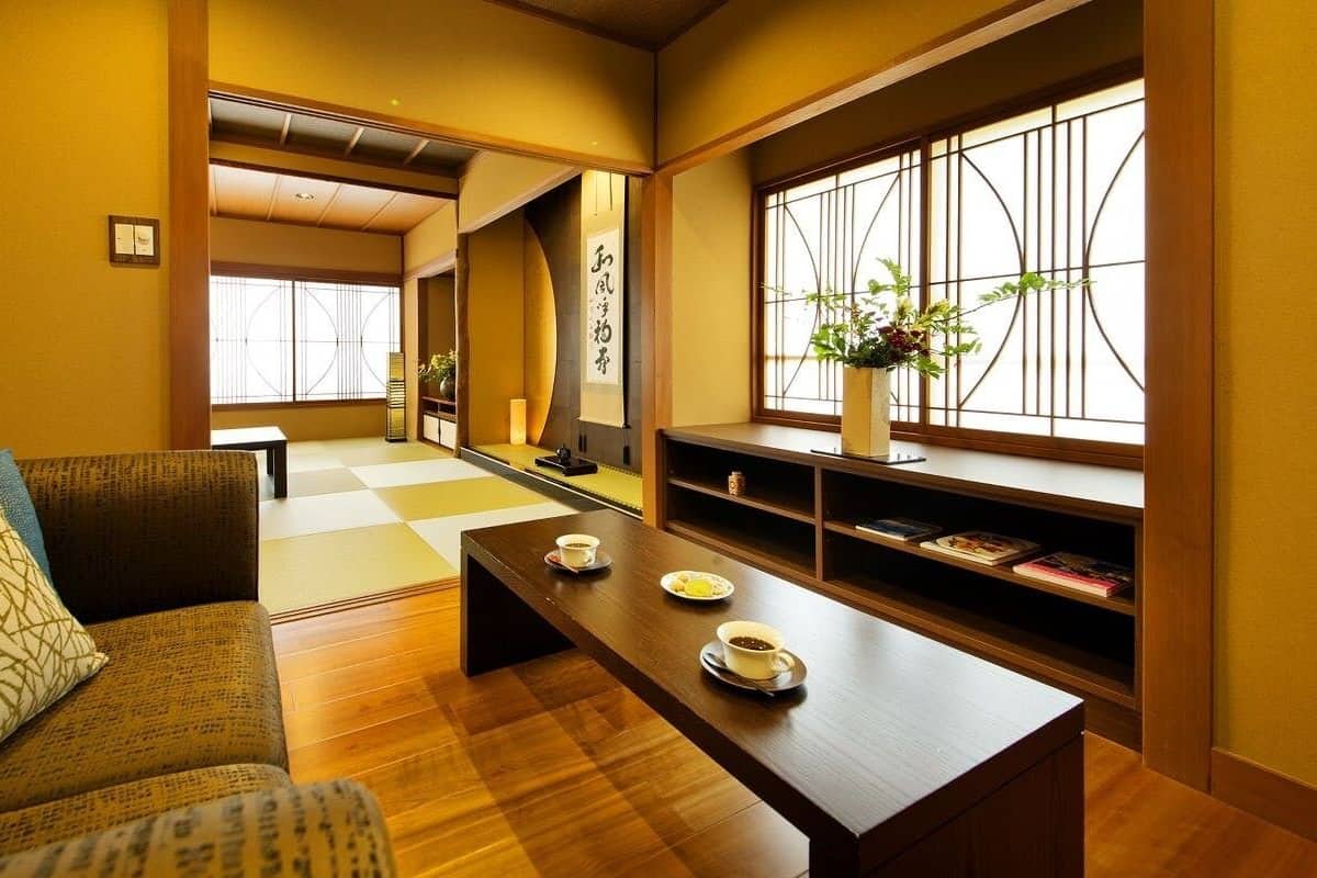 A traditional Japanese room with tatami flooring, a low wooden table with cups, a sofa with cushions, and a window with a geometric pattern.
