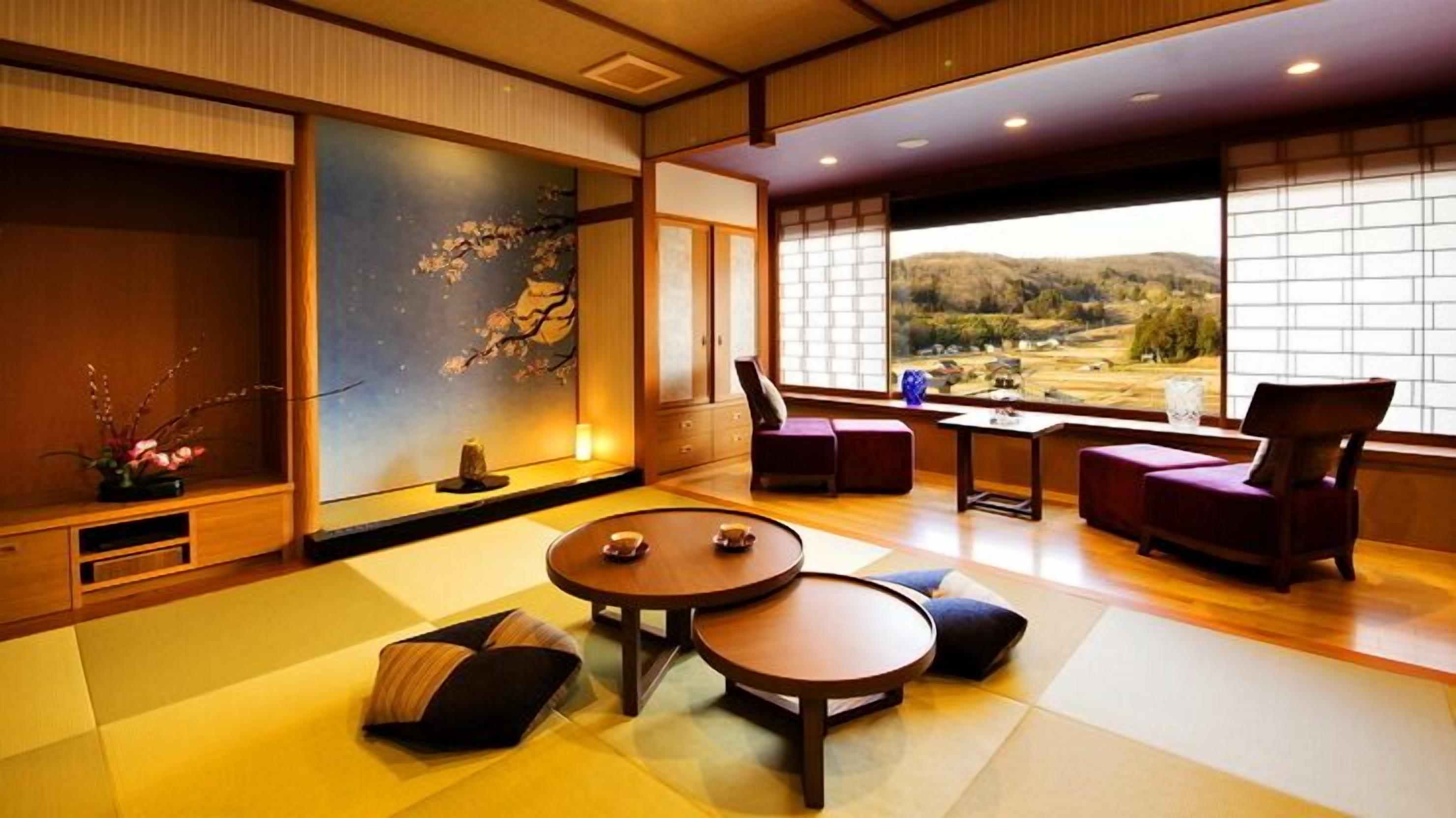Una habitación tradicional japonesa con suelo de tatami, una mesa baja con cojines, vistas a un paisaje montañoso a través de amplios ventanales y un cuadro en la pared.