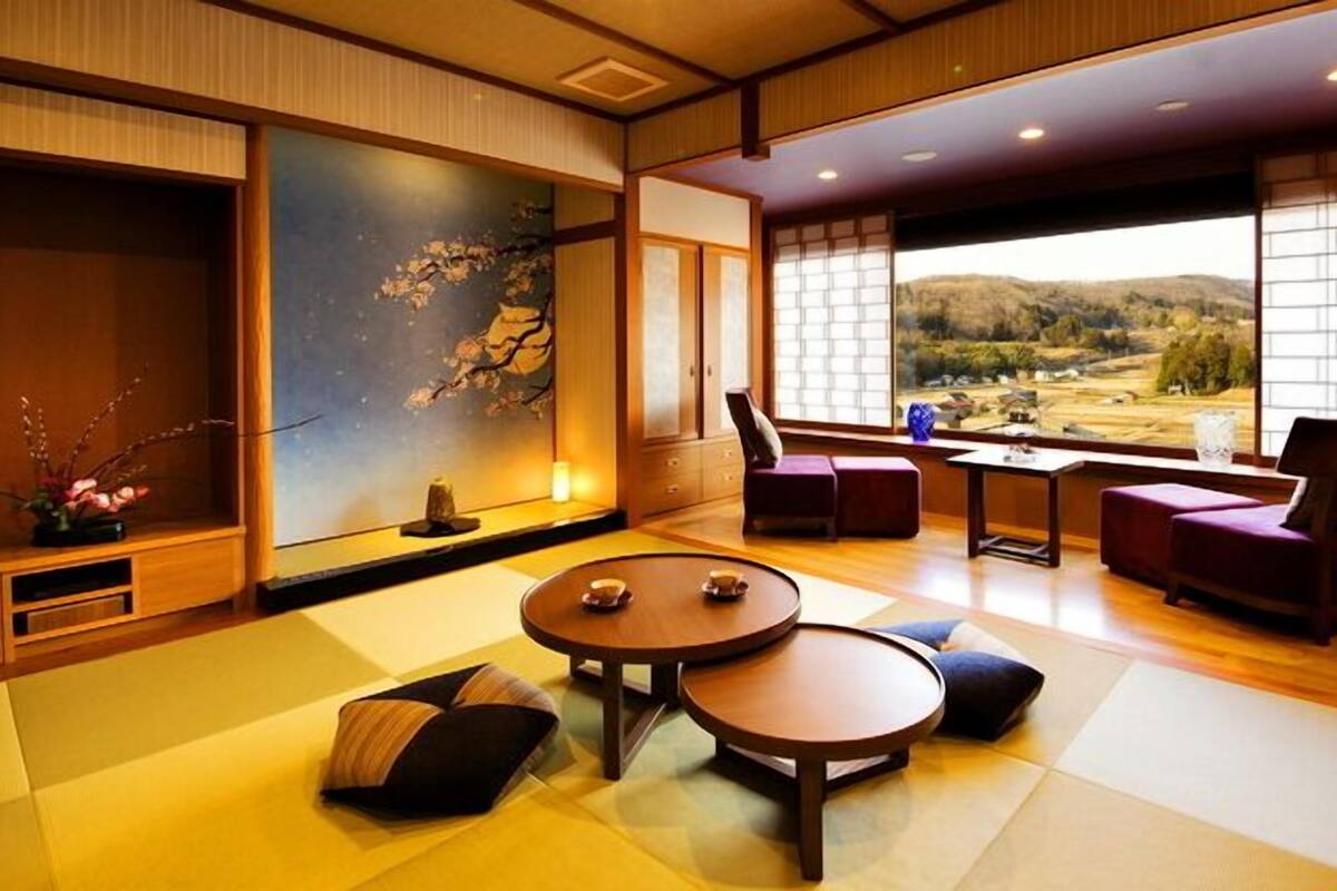 A traditional Japanese-style room with tatami flooring, a low table with cushions, a view of a mountainous landscape through large windows, and a painting on the wall.