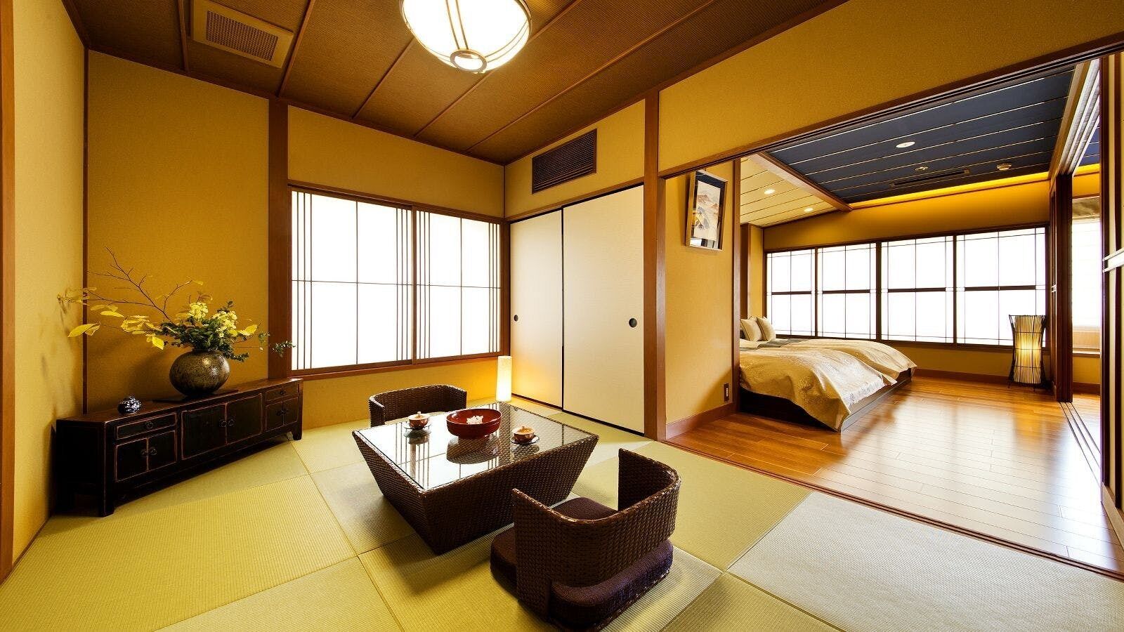 A traditional Japanese room with tatami flooring, a low table with a bowl, a bed with a yellow bedspread, and large windows.