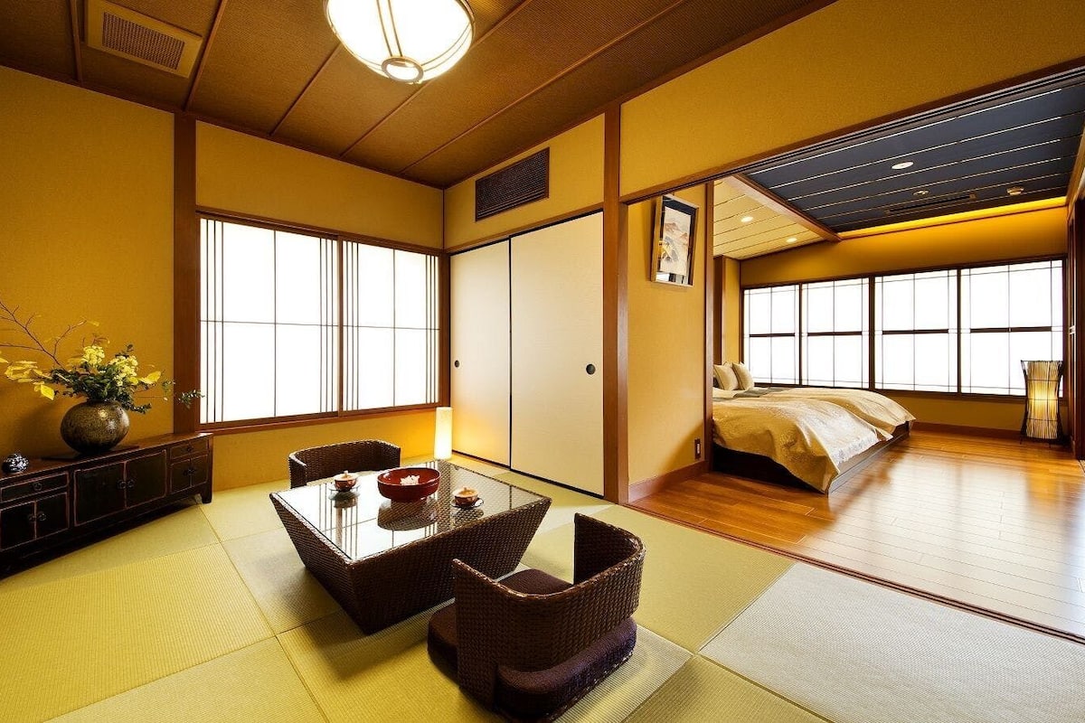 A traditional Japanese room with tatami flooring, a low table with a bowl, a bed with a yellow bedspread, and large windows.