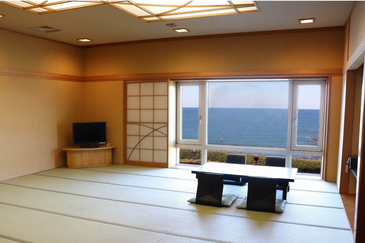 Quarto com piso de tatame, televisão em um suporte, porta deslizante com vista para o mar e uma mesa com cadeiras.