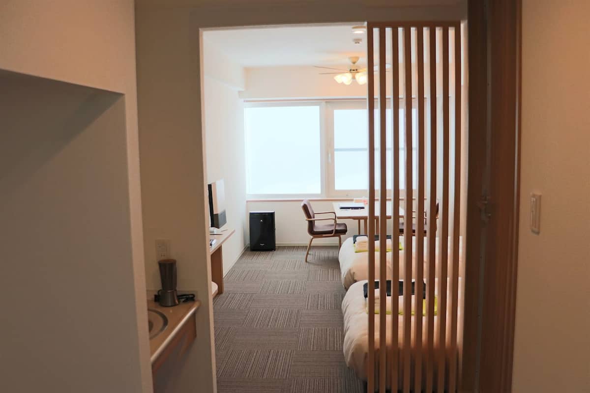 Quarto de hotel com cama, escrivaninha, cadeira, janela e ventoinha no teto.