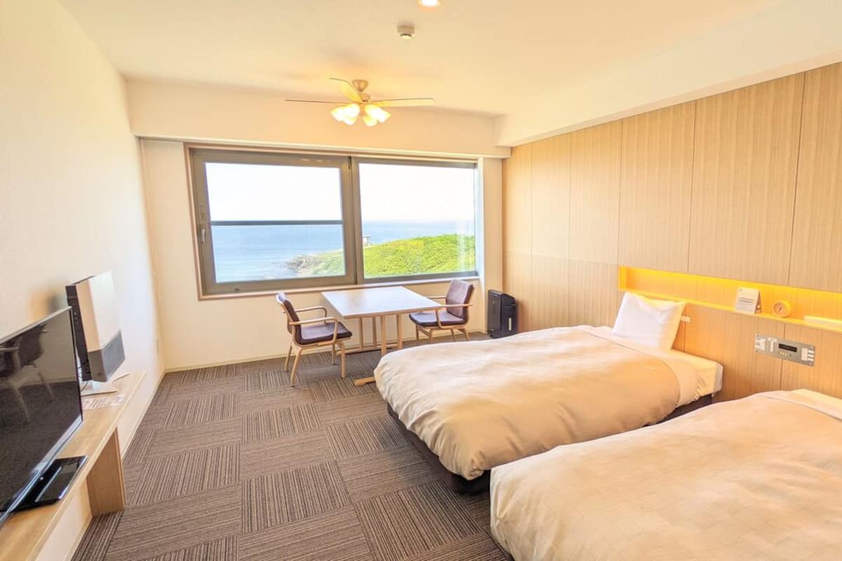 Quarto de hotel com duas camas, uma escrivaninha, uma cadeira, uma televisão e uma grande janela com vista para o mar.