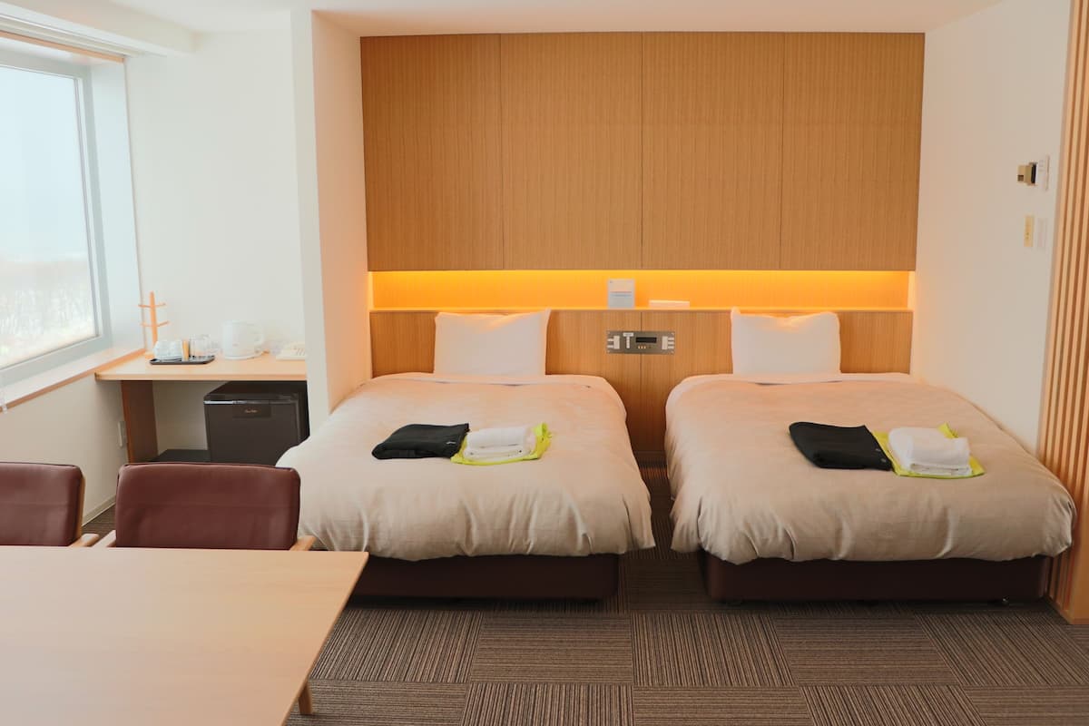 Quarto de hotel com duas camas, uma escrivaninha, uma cadeira e uma janela.