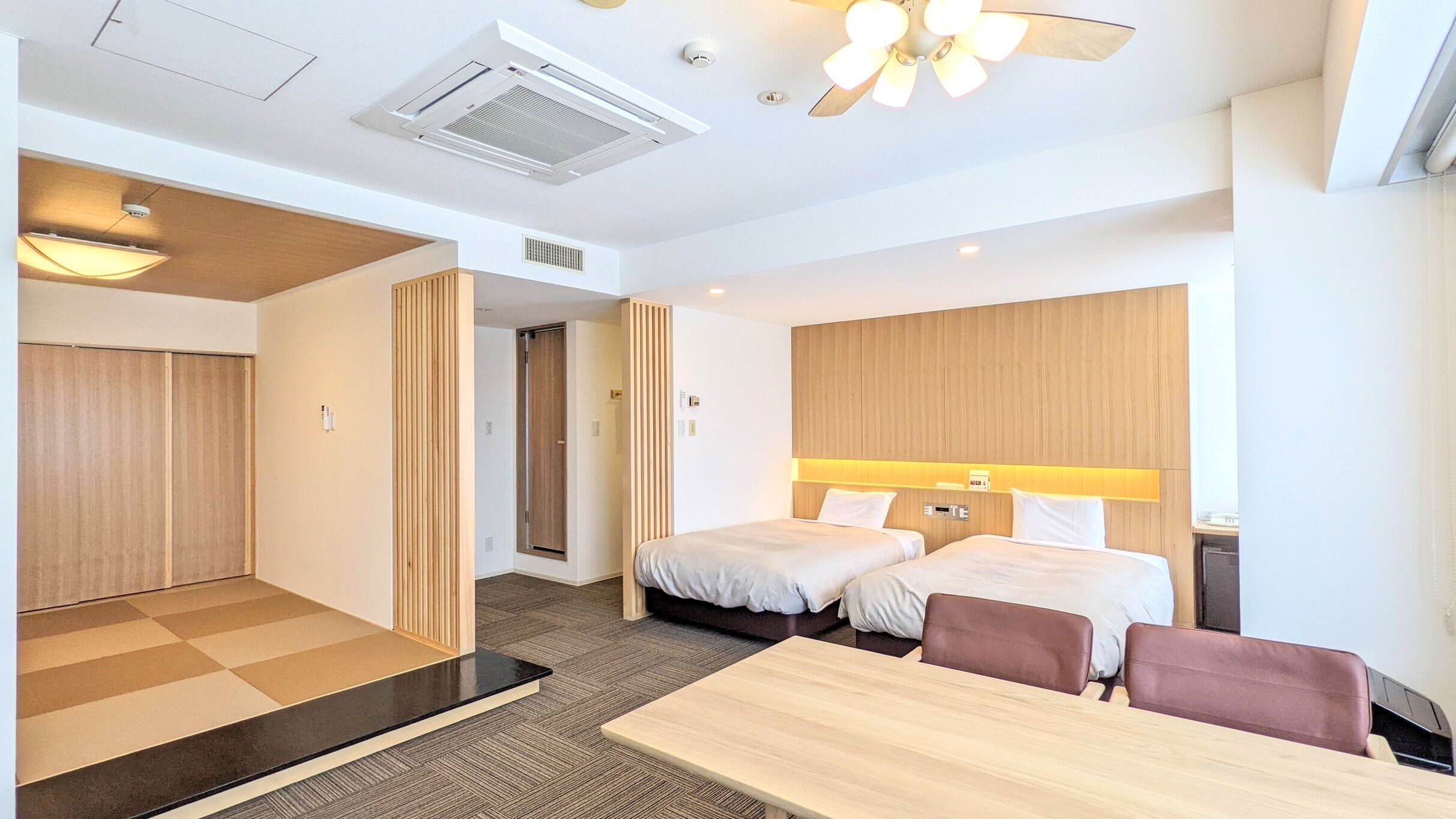 Ein Hotelzimmer mit zwei Betten, einem Schreibtisch, einem Stuhl, einem Deckenventilator und einem Fenster.