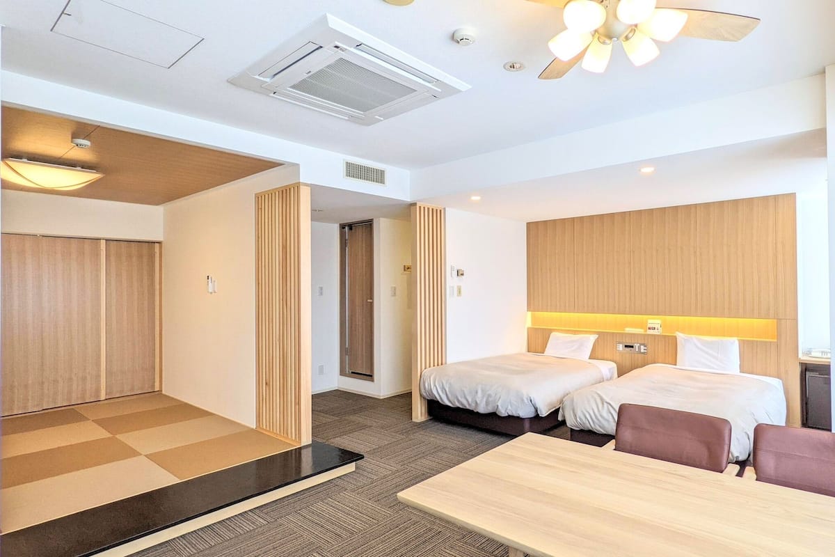 Quarto de hotel com duas camas, uma escrivaninha, uma cadeira, um ventilador de teto e uma janela.