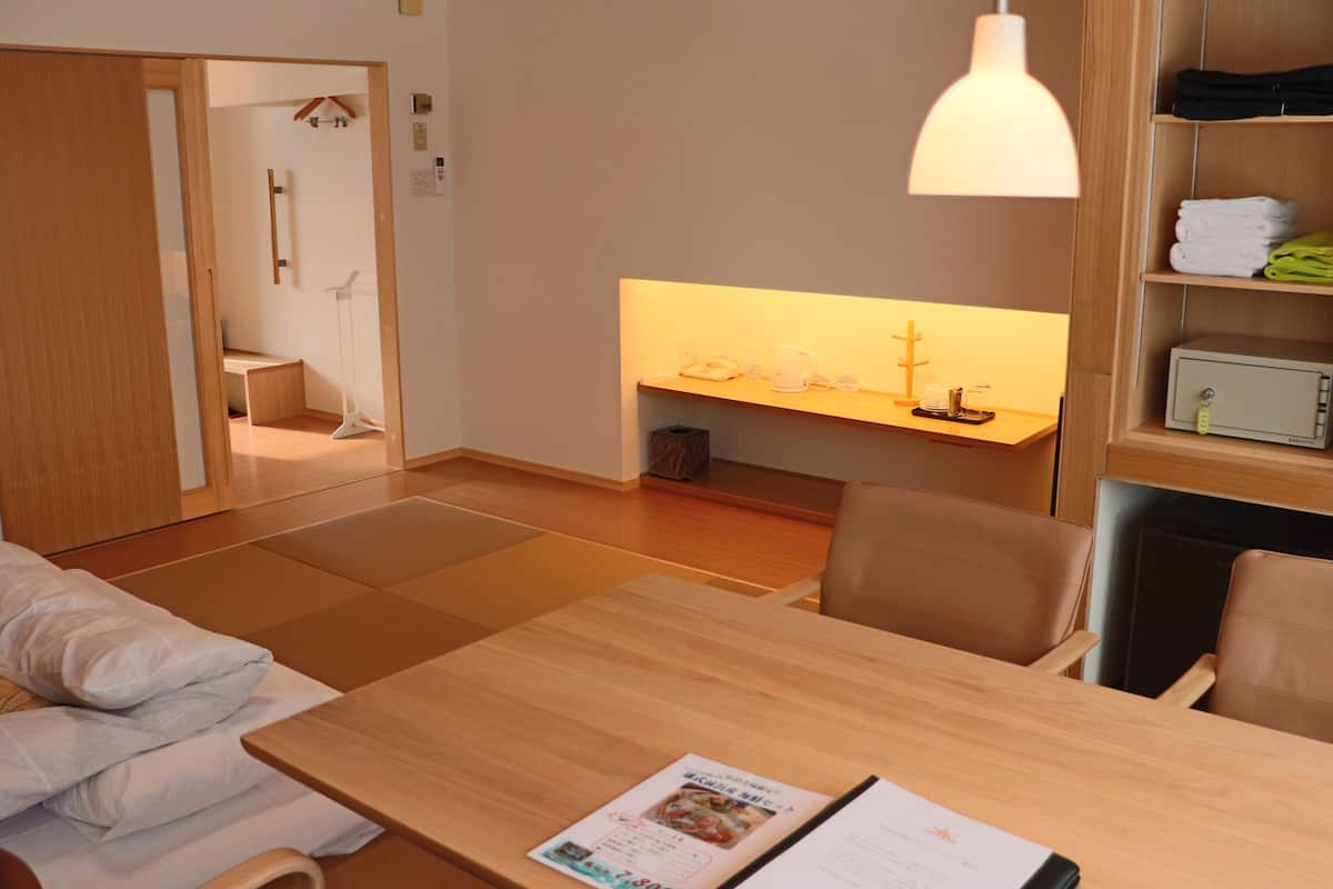 Quarto de hotel com uma mesa de jantar de madeira, luminária pendente, prateleira com toalhas dobradas e vista para outro quarto.