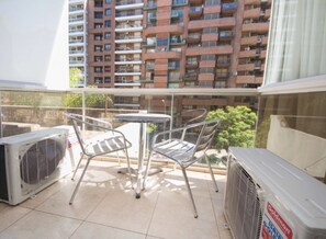 Uma varanda com mesa e cadeiras, aparelhos de ar condicionado e vista para um edifício alto com várias varandas.
