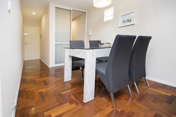 Uma área de refeições com mesa de vidro e quatro cadeiras cinzas, piso de madeira, porta branca e um quadro na parede.