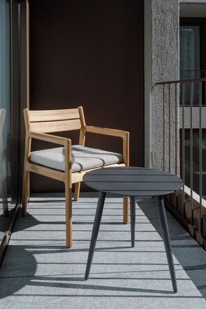 A wooden chair with a cushion and a small table on a balcony with a concrete floor and a metal railing.