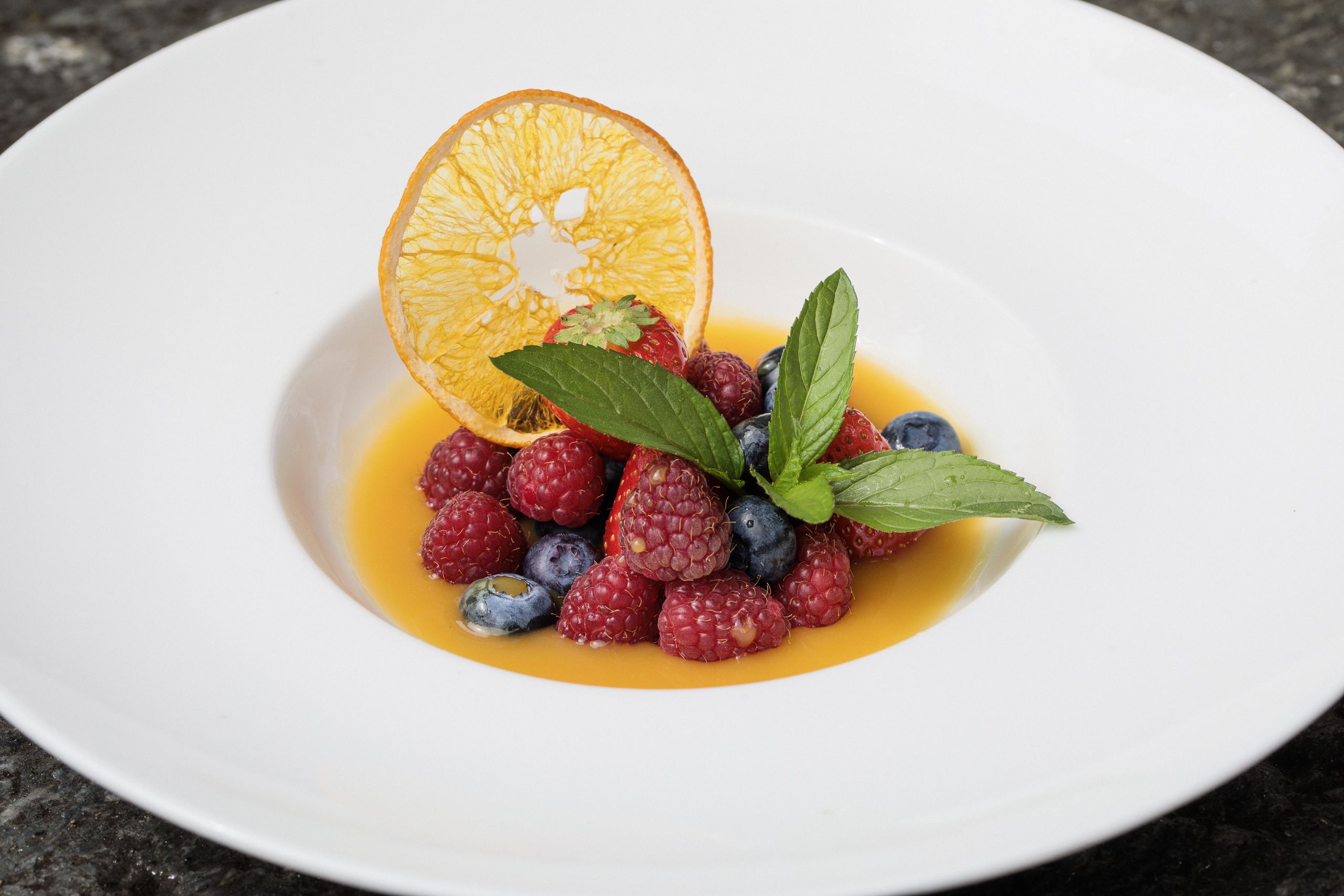 Ein weißer Teller mit einem Dessert aus Himbeeren, Blaubeeren und einer Orangenscheibe, garniert mit Minzblättern.