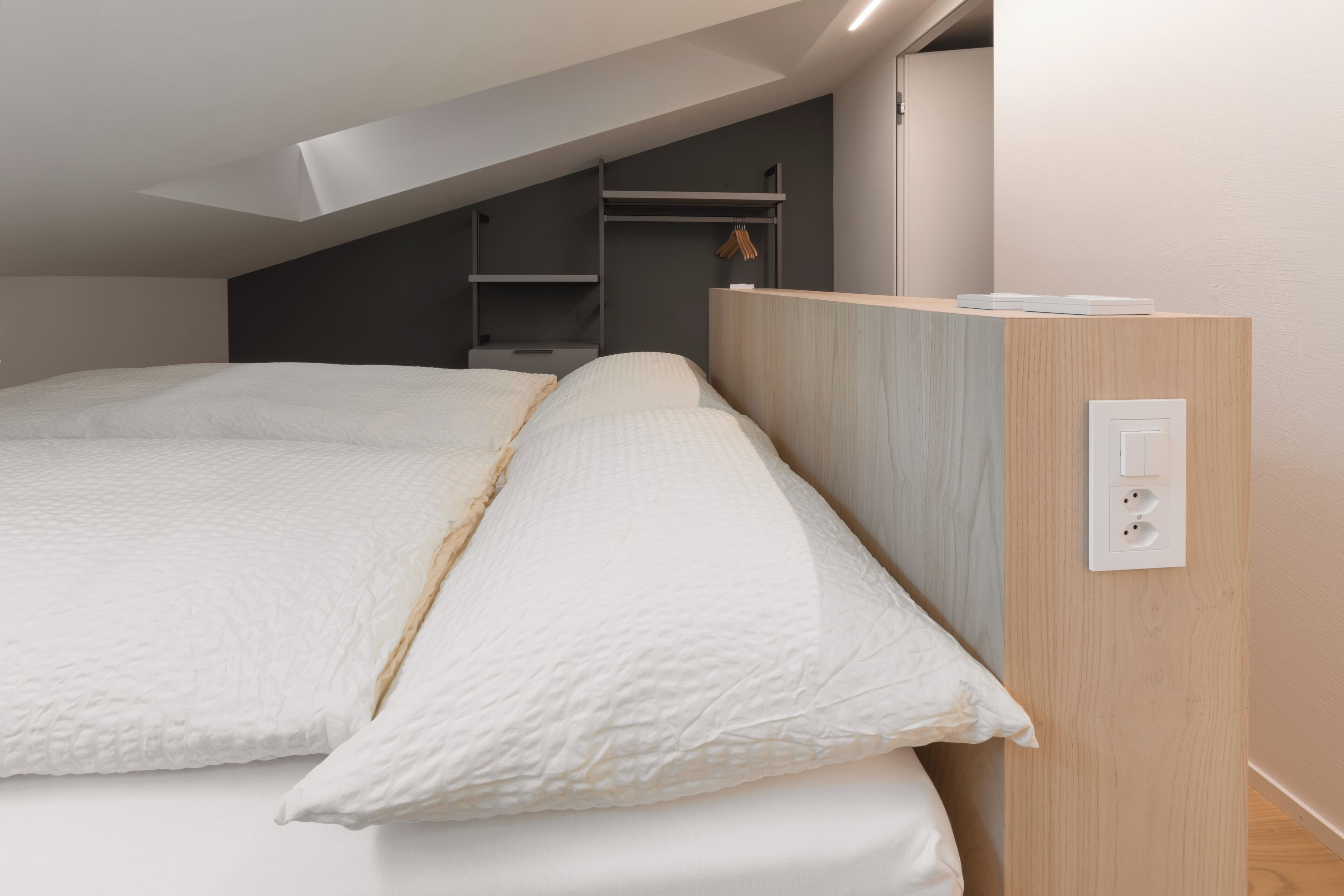 Un dormitorio con techo inclinado, una cama con ropa de cama blanca, una mesita de noche de madera y un enchufe en la pared.