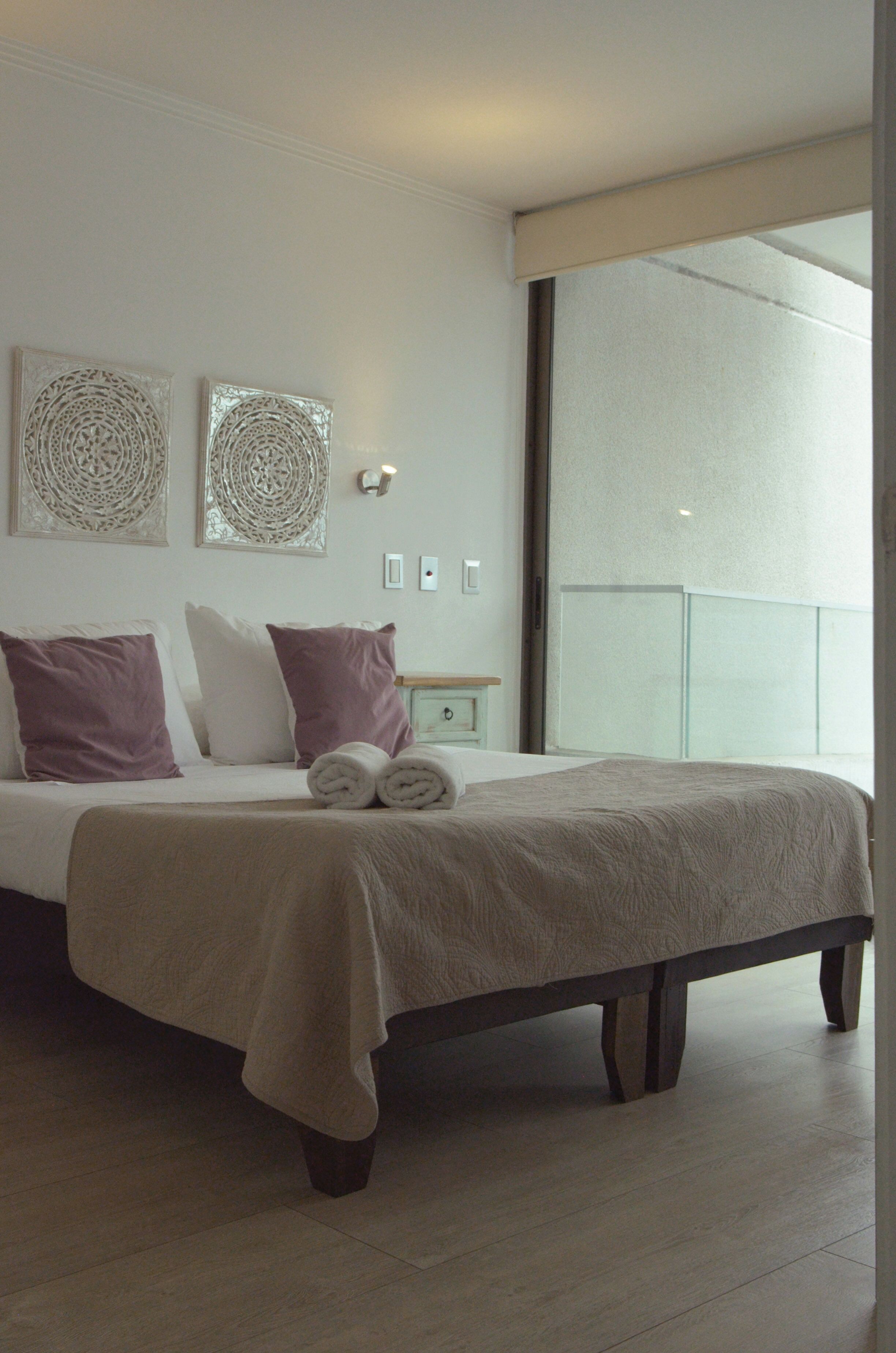 A modern bedroom with a large bed, two decorative wall pieces, a nightstand, and a large window.