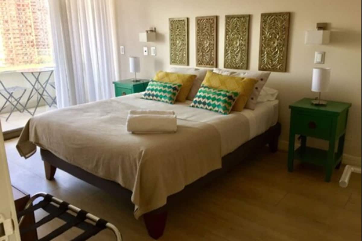 Ein ordentlich eingerichtetes Schlafzimmer mit einem Bett, Kissen, einem grünen Nachttisch und einem Fenster mit Vorhängen.