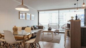 A modern living room with a glass dining table, a sofa, and a kitchen area with a wooden cabinet. - Loica Suites (Santiago)