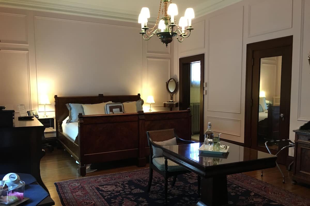 A hotel room with a wooden bed, a glass table, a chair, a desk, and a desk lamp.