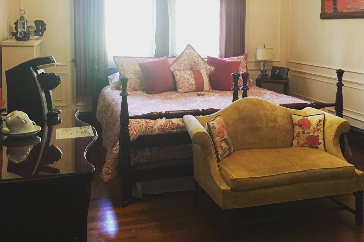 A bedroom with a bed, a chair, a desk, and a painting on the wall.