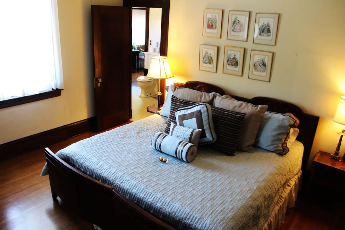 A neatly made bed with a striped bedspread and several pillows, a bedside table with a lamp, and a wall with framed pictures.