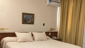 A hotel room with a bed, a nightstand, a chair, a painting on the wall, and an air conditioning unit. - Hotel Cervantes (Mendoza)