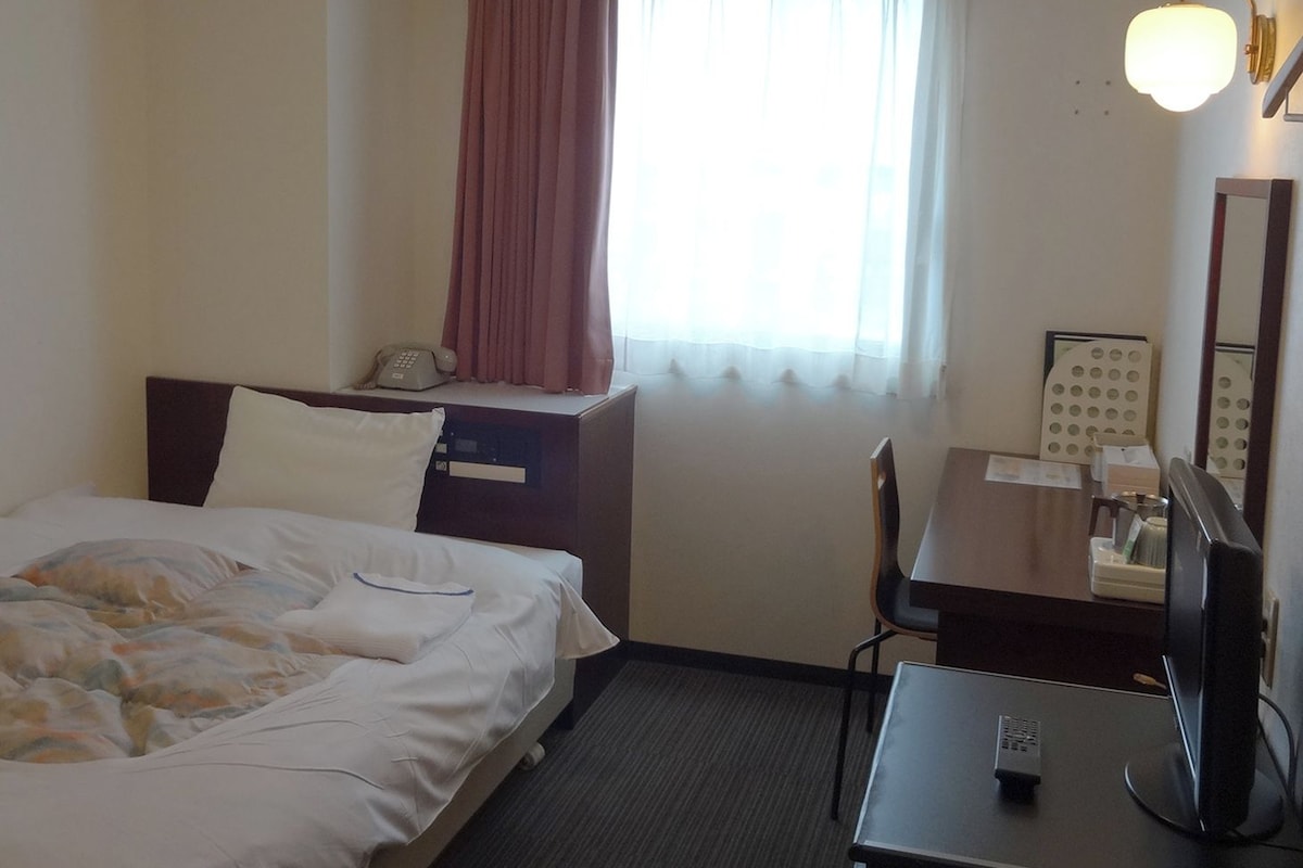 A hotel room with a bed, a desk, a chair, a telephone, a television, and a window with curtains.