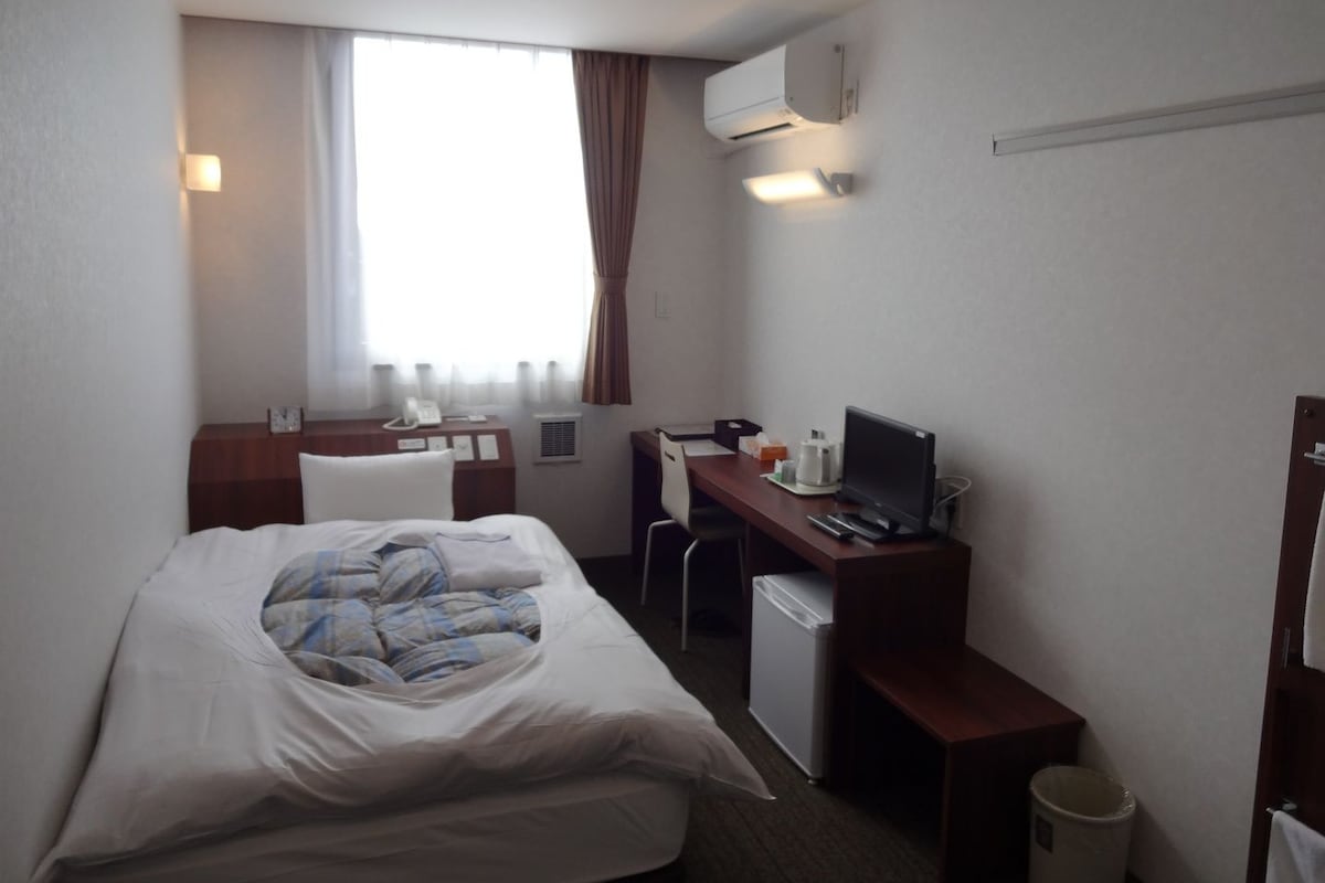 A hotel room with a bed, a desk with a computer, a chair, a small fridge, and an air conditioning unit.