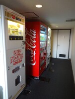 Uma área com máquinas de venda automática, incluindo uma máquina de Coca-Cola e uma de Cup Noodle.