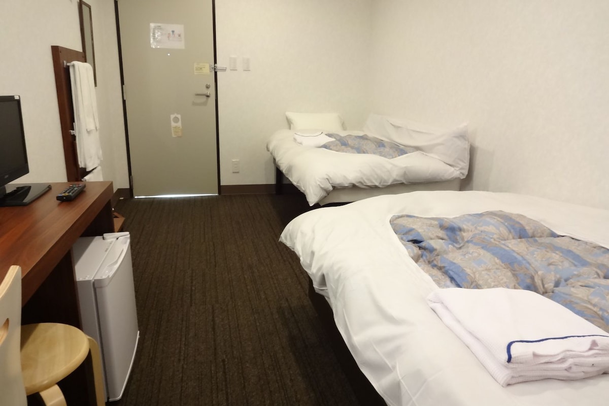 A hotel room with two beds, a desk with a computer and a remote control, a chair, and a small trash bin.