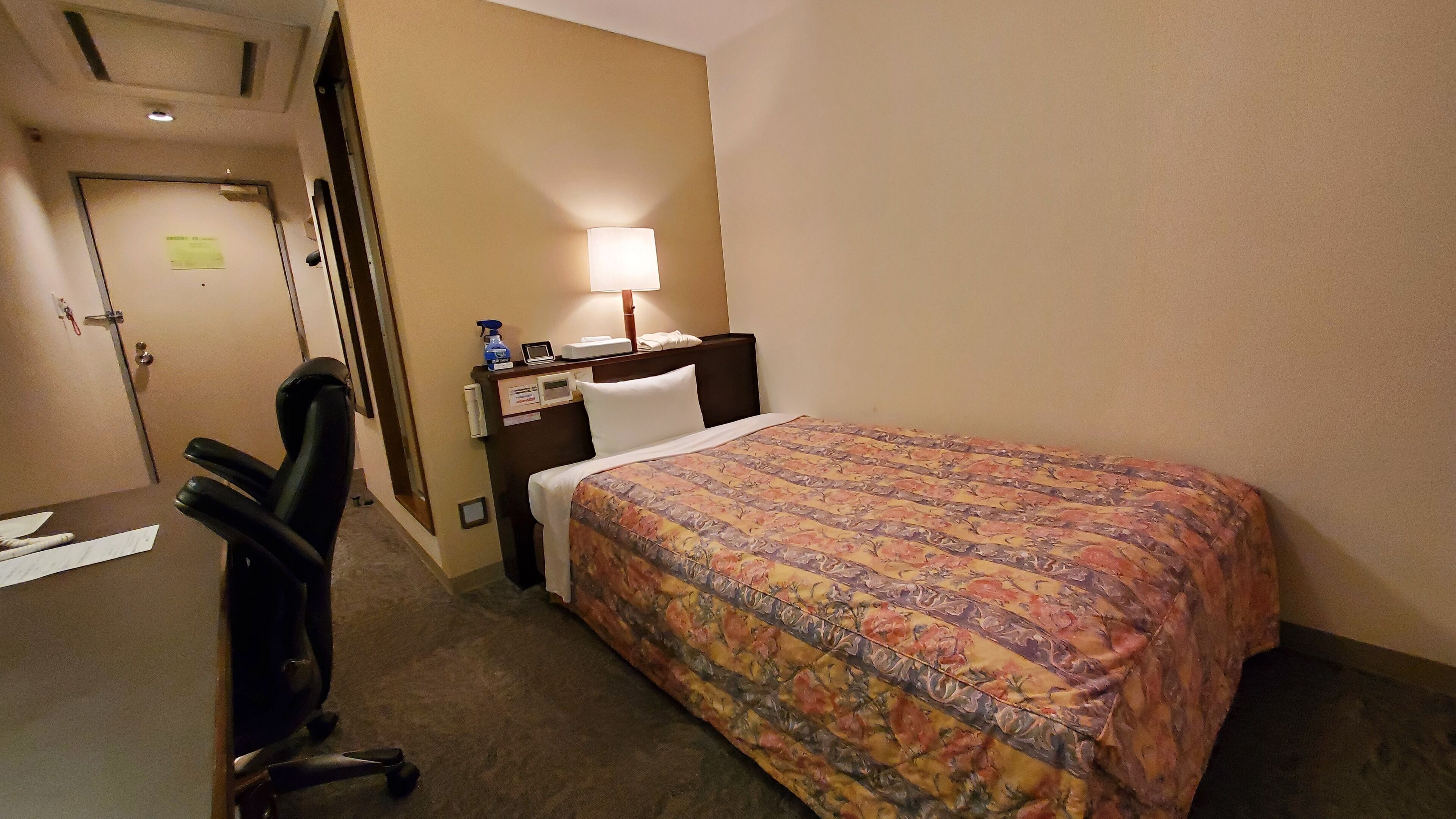 Habitació d'hotel amb un llit, un escriptori, una cadira, una làmpada i una porta.